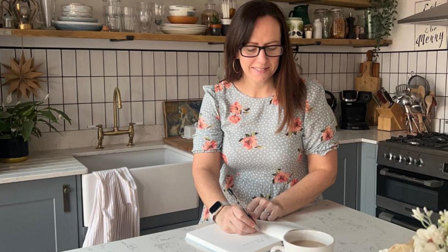 Woman sitting at a kitchen island with a white, marble, quartz worktop. A Belfast sink is behind, along with oak shelves.