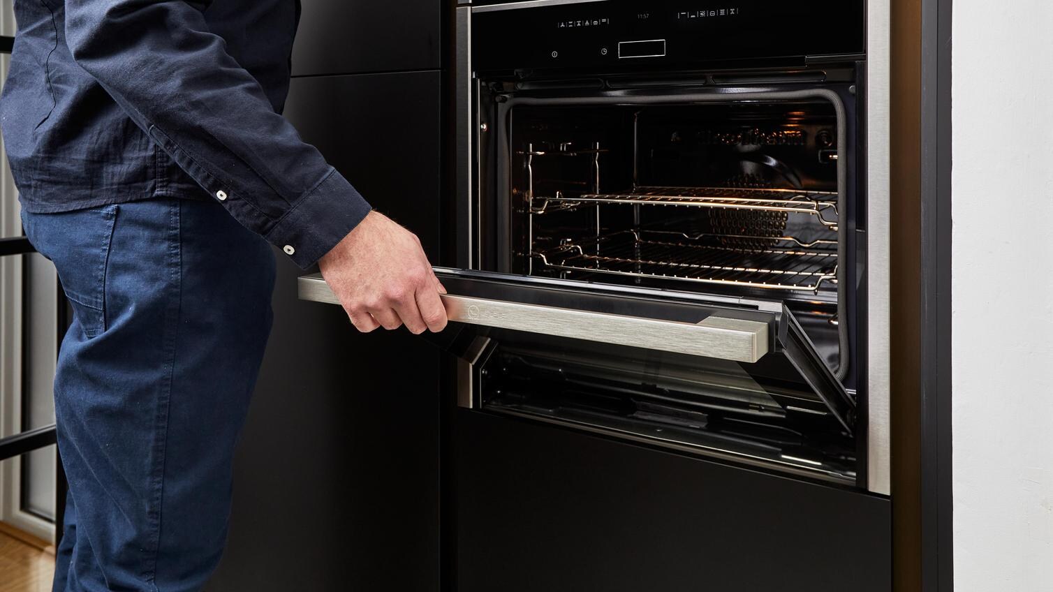 Man wearing blue shirt and jeans opening an integrated, black oven in a tower cabinet.