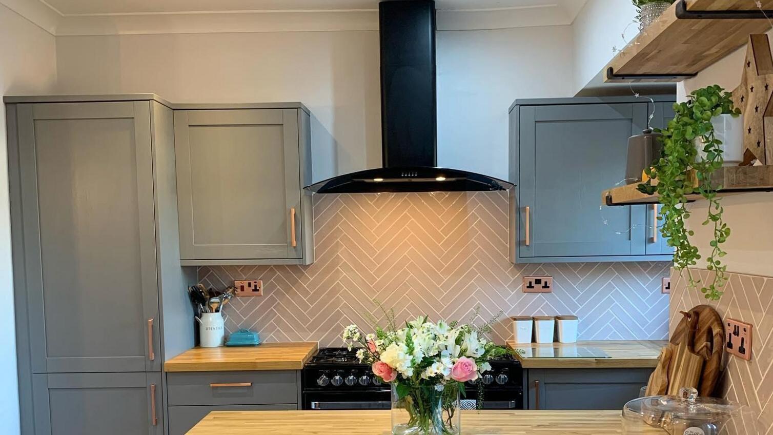 Dark-grey kitchen with shaker cupboard doors and a breakfast bar. Has copper handles , wood worktops, and a black extractor.