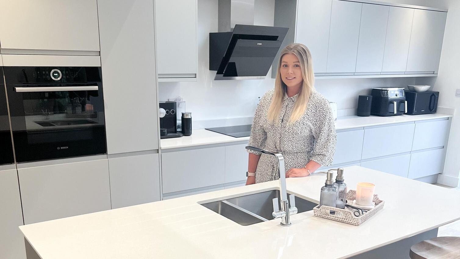 A calm haven kitchen makeover with grey integrated handle doors in an island layout. Has white worktops and a black oven.