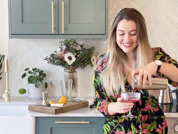 Poppy stood in front of a teal shaker kitchen, wearing a floral dress, looking down, and pouring a cocktail into a glass.