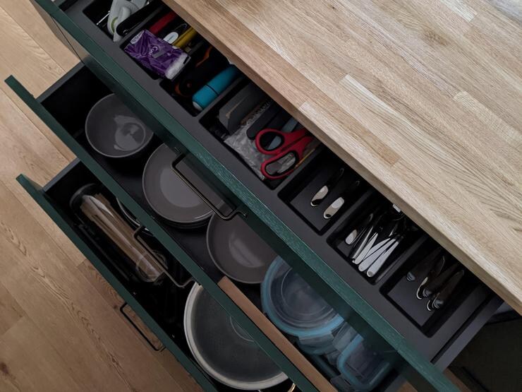 Birds eye view of three green shaker doors open, showcasing cutlery, bowls, and kitchenware. With oak floors and worktop.