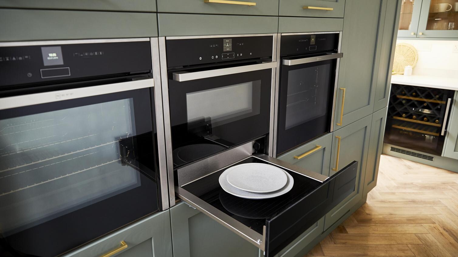 Sage green shaker kitchen with tower wall units, featuring three built in ovens in a row, with a warming drawer pulled out.