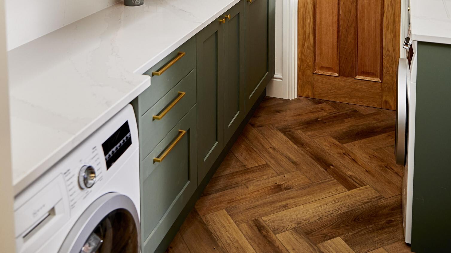 Sage green shaker utility room design with white quartz worktop, brass bar handles, a washing machine, and an oak door.