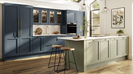 A two-tone kitchen with blue and green shaker doors, white worktops, wooden floors, and brass knobs in a peninsula layout