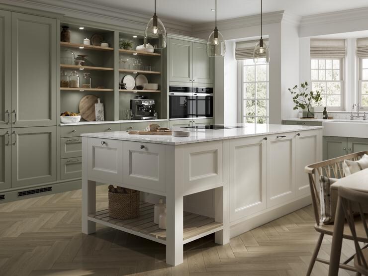 A sage-green shaker kitchen idea with an ivory island. Has white worktops, open shelving, chevron floors, and brass handles.