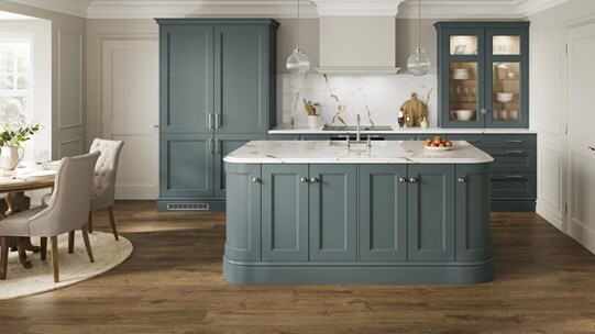A light blue shaker kitchen in a single wall and island layout. It is finished with white worktops and oak flooring