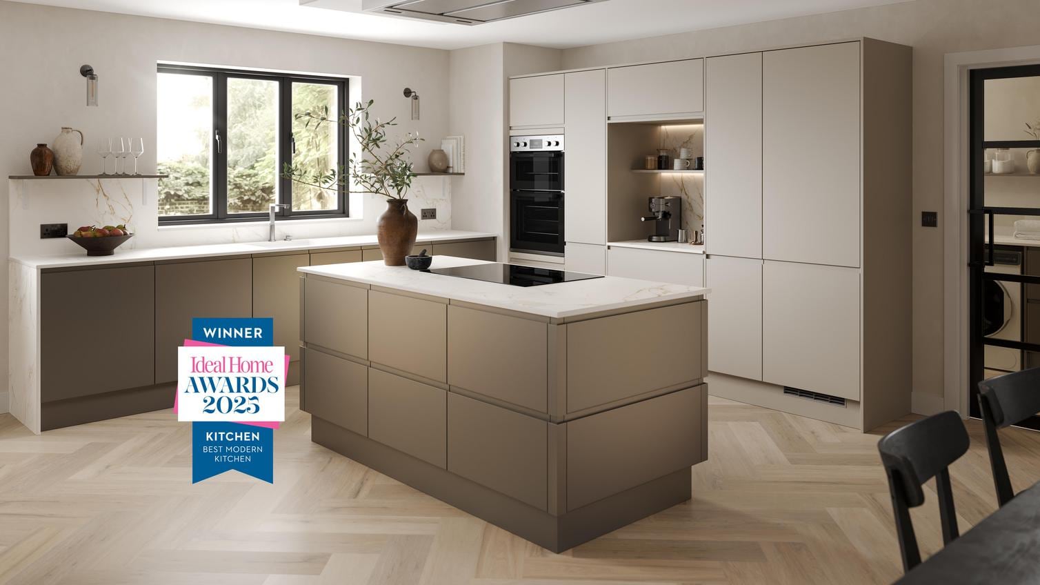 A handleless, bronze kitchen in an L-shaped, island layout. It has white worktops, chevron flooring, and an induction hob.