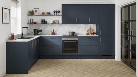 A marine blue kitchen with slab doors and brass handles. It is in an l-shaped layout, with grey worktop and oak flooring.