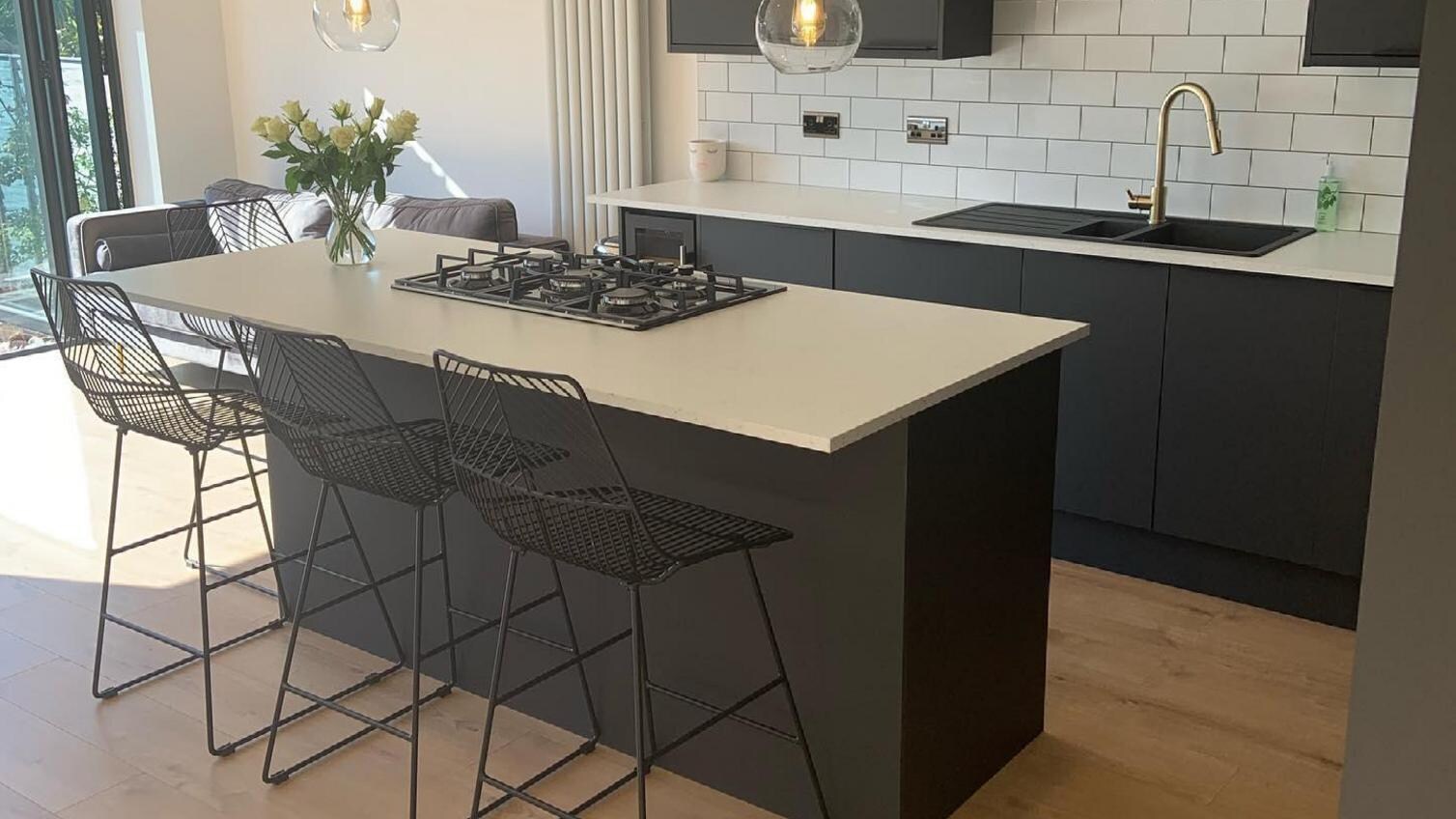 Black integrated handle kitchen with an island, black stools, a white worktop and gold tap.