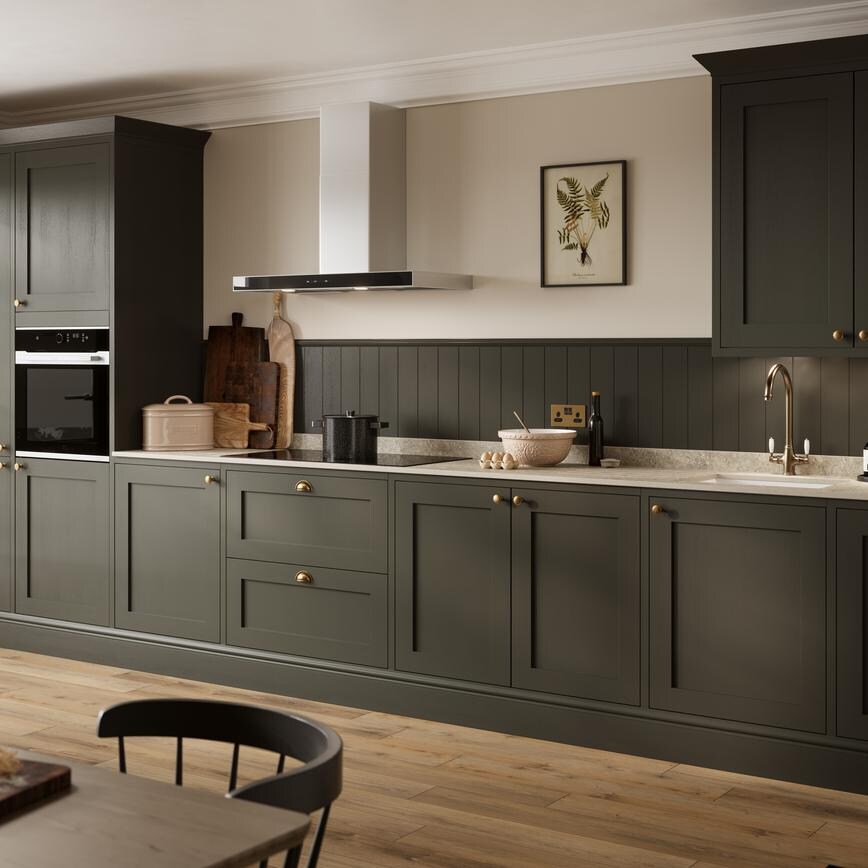 A dark-green kitchen with shaker doors in a single wall layout. Styled with brass handles, white worktops, and oak flooring.
