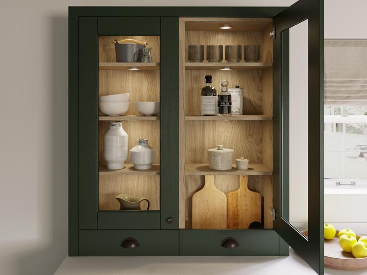 Dark-green kitchen cupboard with a glass door front, showing wooden shelves and neutral crockery, above a white worktop.