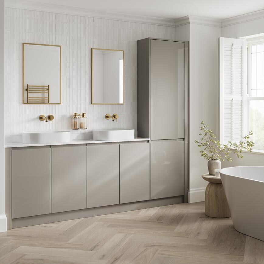 A bright bathroom with gloss, sand grey cabinets. It has oak-effect flooring, brass fixtures, and a large roll-top bath. 